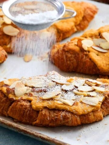 a hand dusting white sugar over pumpkin almond croissants.