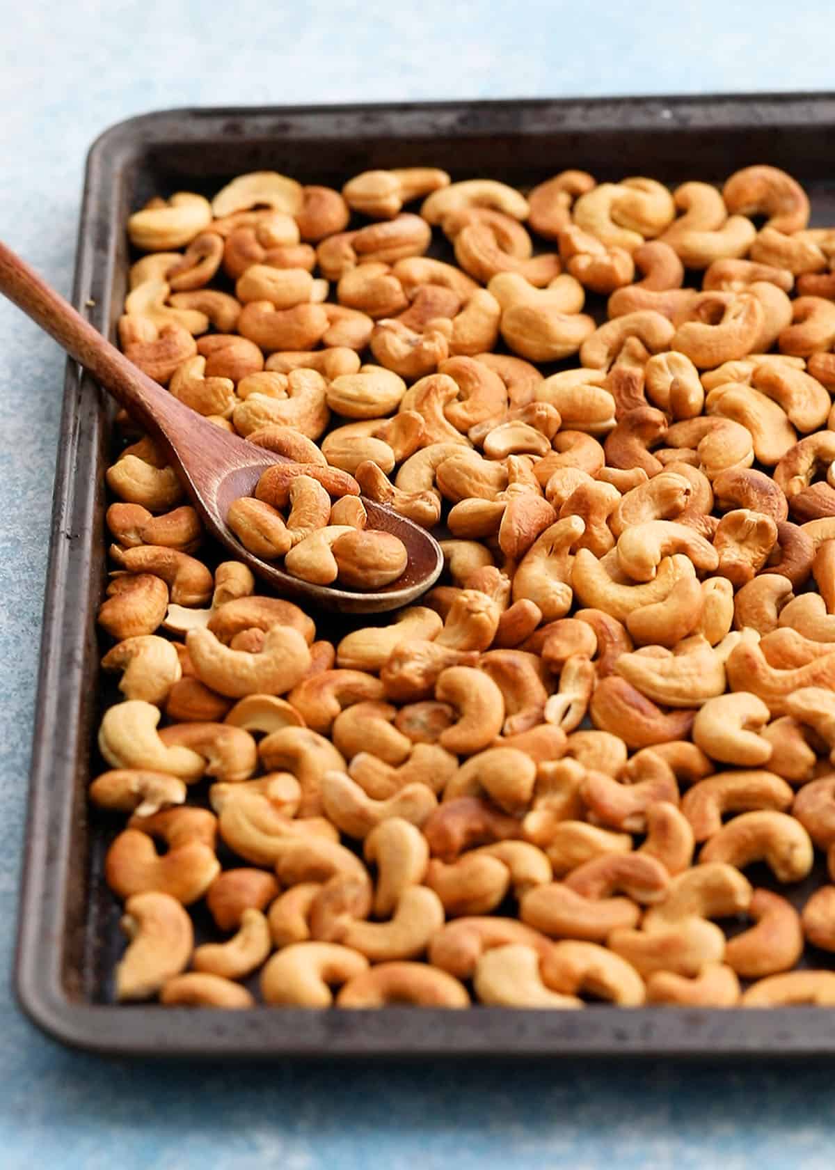roasted cashews in a metal baking sheet along with wooden spoon.