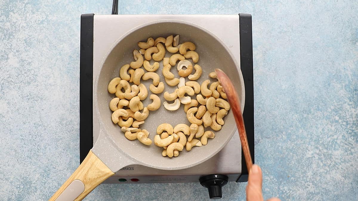 a hand stirring raw cashews in a small white skillet.