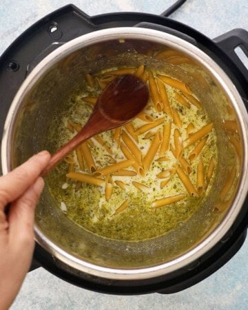 a hand mixing raw penne pasta along with a green liquid in an instant pot.