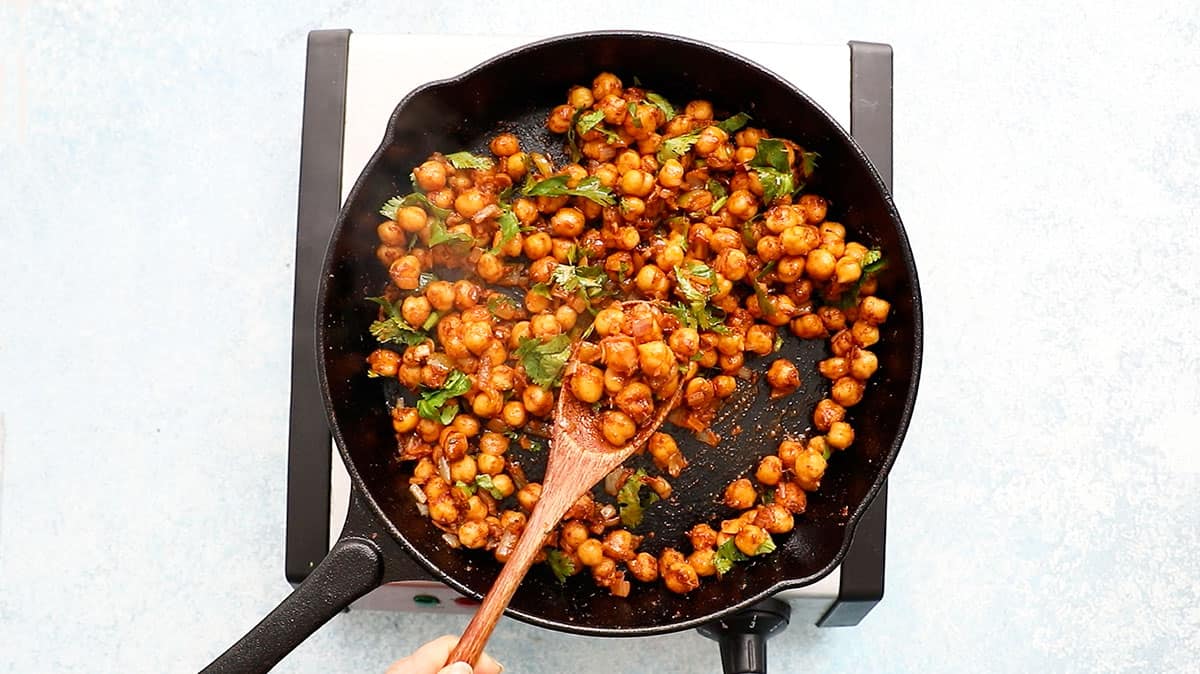 a hand lifting cooked chickpeas from a black skillet using a wooden spoon.