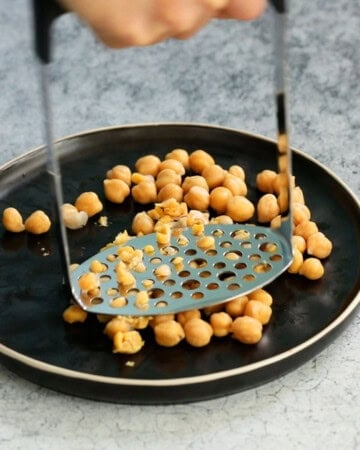 a hand mashing chickpeas placed on a black plate using a potato masher.
