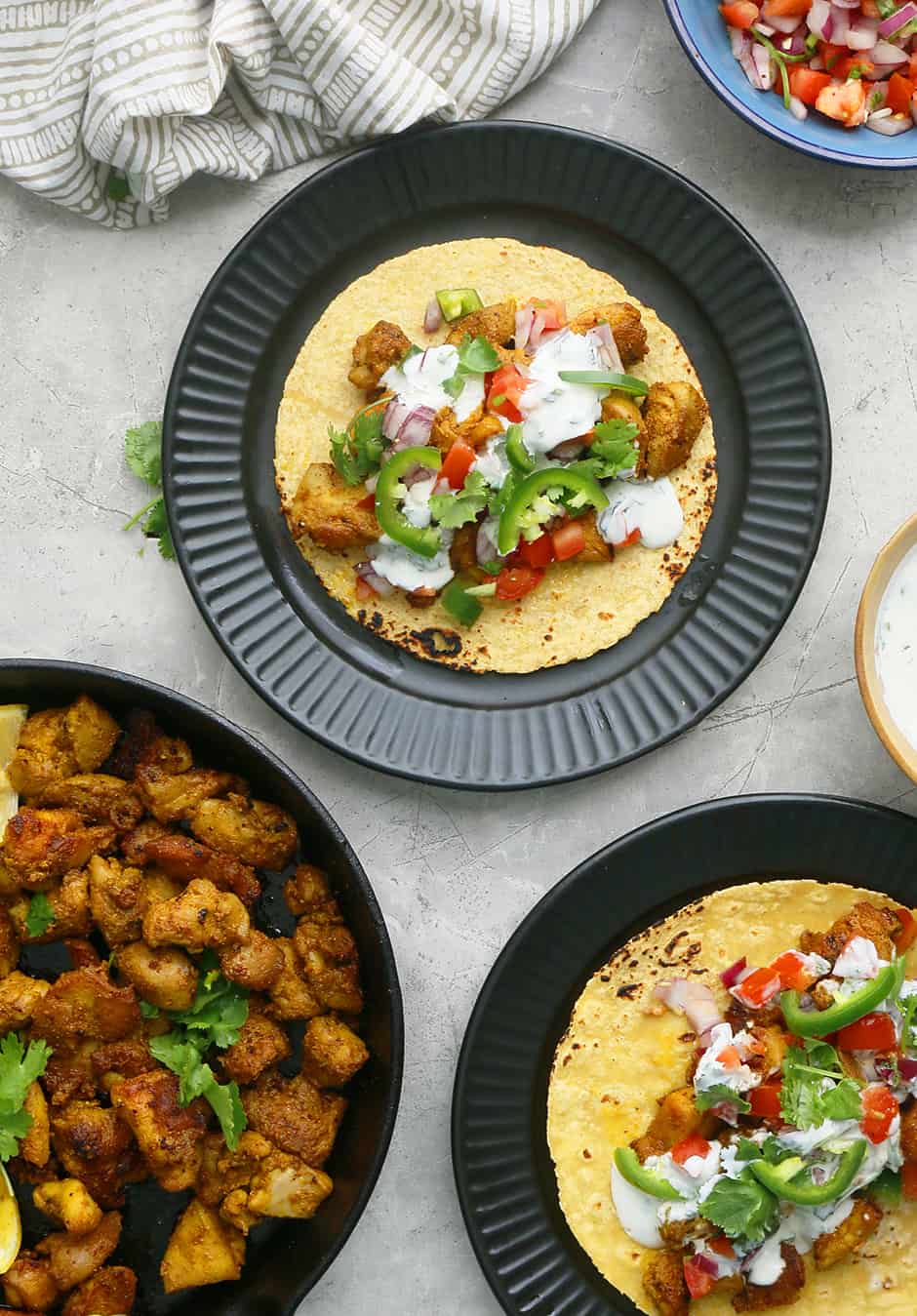 2 black plates with chicken tacos, a blue bowl with pico de gallo and more spicy chicken