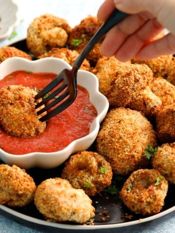 a hand dipping one breaded mushroom in red sauce placed in a white bowl.