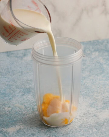 a hand pouring white milk over yellow mango chunks placed in a blender.