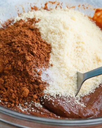 a hand mixing white flour, brown cocoa powder and melted chocolate in a glass bowl.