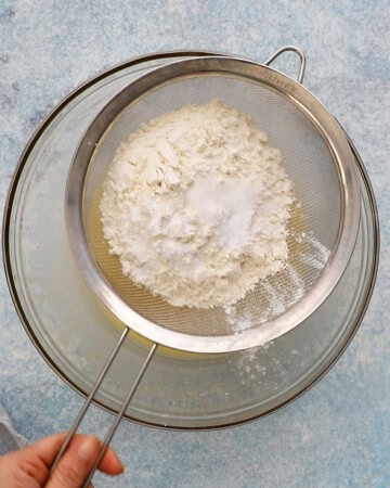 a hand sifting white flour over a glass bowl