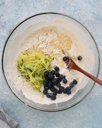grated zucchini and blueberries along with muffin batter in a large glass bowl.