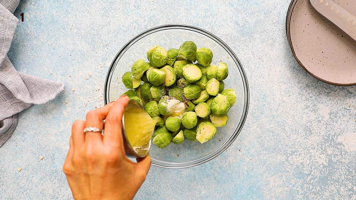 a hand pouring melted butter into a glass bowl filled with brussels sprouts.