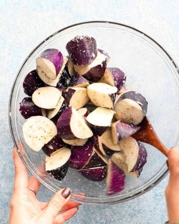 two hands tossing eggplant chunks in a glass bowl.