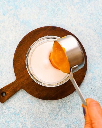 a hand adding yellow mango puree into white milk placed in a glass jar.