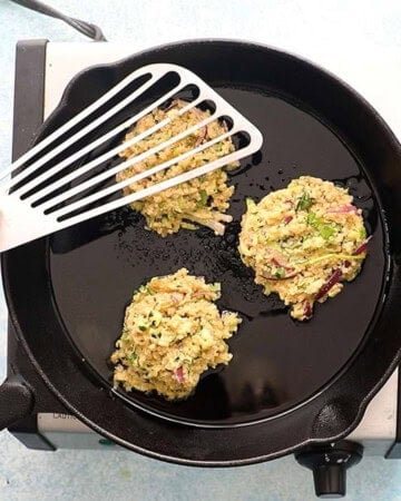 three zucchini fritters cooking in a black skillet.