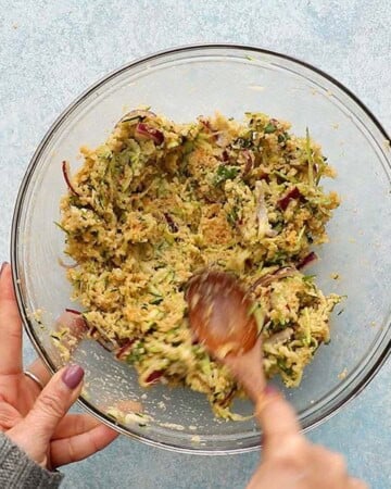two hands mixing zucchini fritters mixture in a glass bowl.