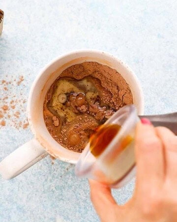 a hand pouring brown vanilla extract into a white mug.