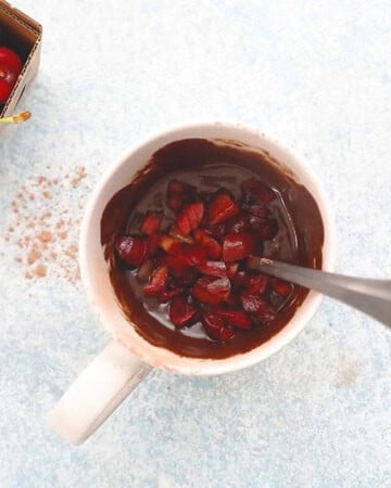 one white mug with brown chocolate cake batter topped with chopped red cherries.