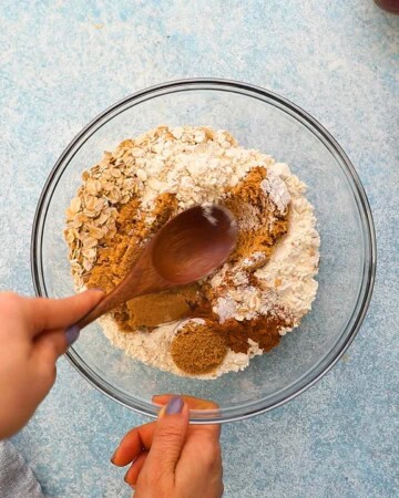 two hands mixing white flour along with brown sugar in a glass bowl.