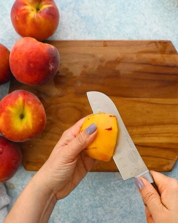 two hands cutting one yellow peach into slices using a knife.