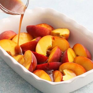 brown liquid being poured into a white dish filled with sliced yellow peaches.