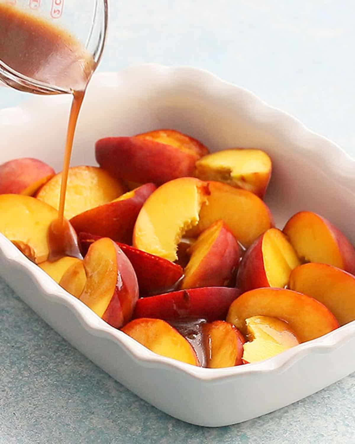 brown liquid being poured into a white baking dish with sliced peaches.