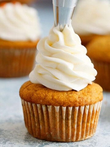 a hand piping white frosting on top of a pumpkin cupcake.