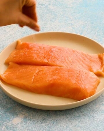 a hand seasoning two salmon fillets placed on a white plate.