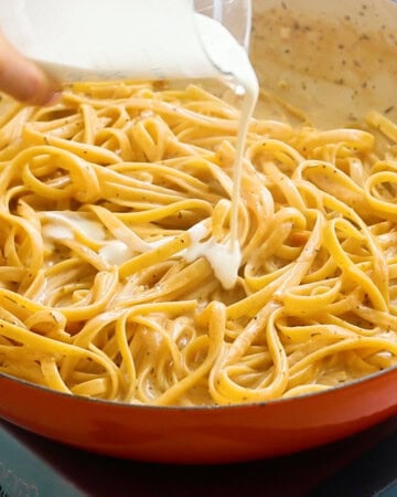 a hand pouring white cream over cooked fettuccini.