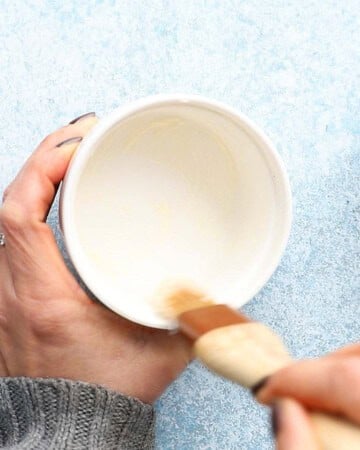 two hands brushing melted butter in a small white ramekin.