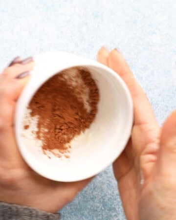 two hands dusting one white white ramekin with brown cocoa powder.