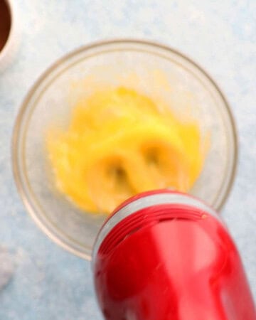 a hand beating eggs in a glass bowl using a red beater.