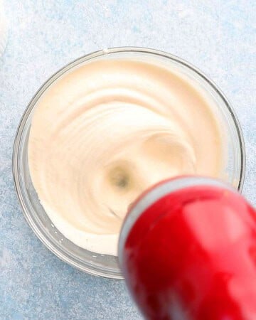 a hand beating eggs in a glass bowl using a red beater.