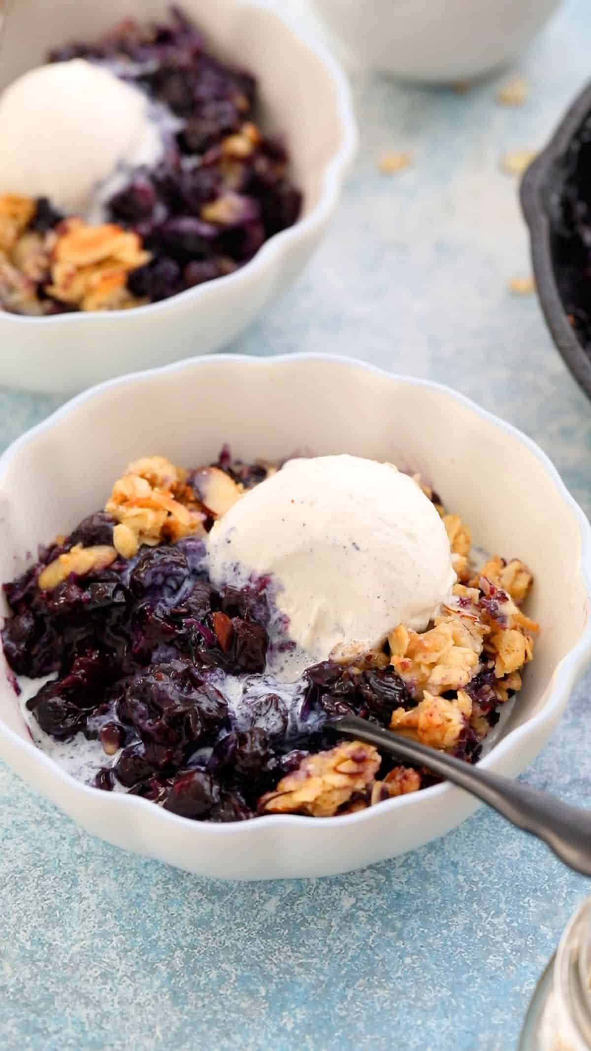 two white bowls with blueberry crisp with crumble topping and one scoop of white ice cream.