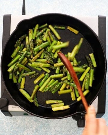 a hand sauteeing chopped green asparagus in a black skillet.