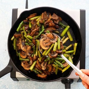 a hand lifting a ladle full of cooked mushroom and asparagus.