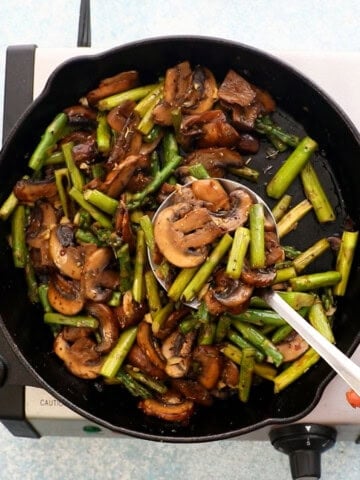 a hand lifting a ladle full of cooked mushroom and asparagus.
