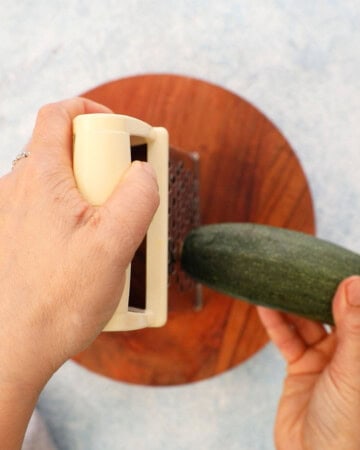 two hands grating one green zucchini using a box grater.