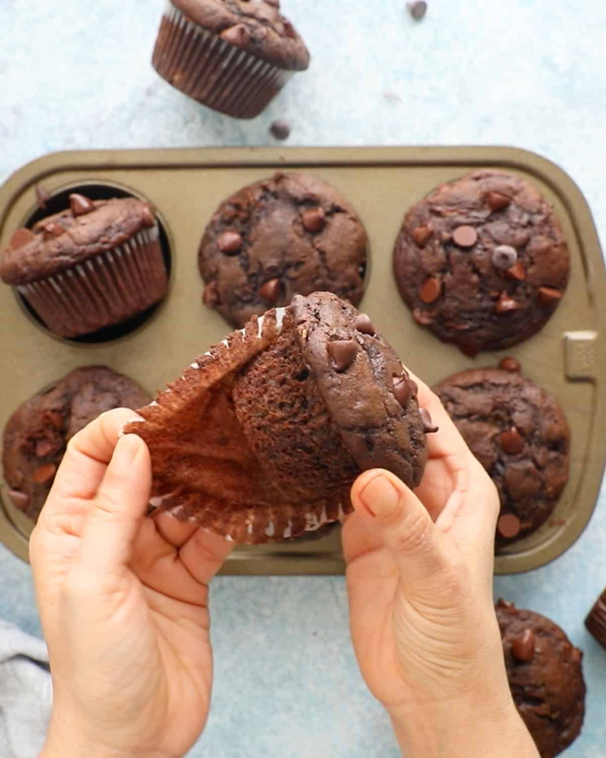 two hands peeling a paper liner from one chocolate muffin .