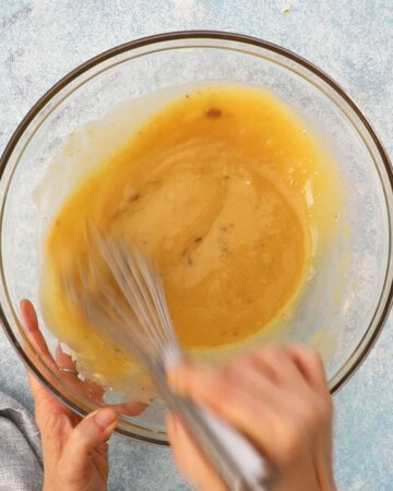 two hands whisking wet ingredients in a large glass bowl.