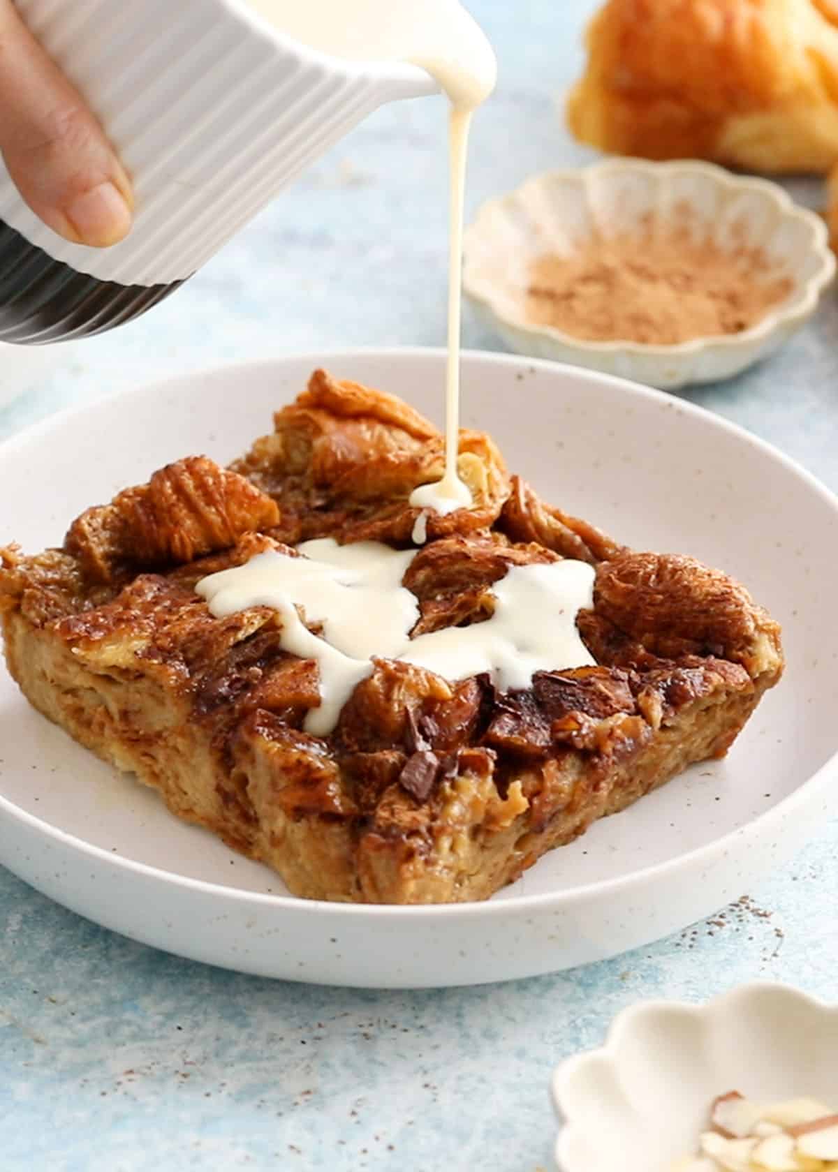 a hand pouring white sauce over a square piece of croissant bread pudding placed on a white plate.