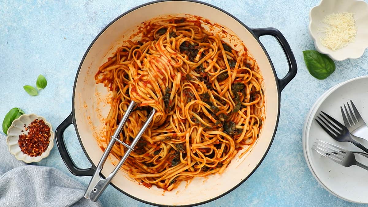 cooked red sauce pasta in a large white skillet along with a metal tongs. 