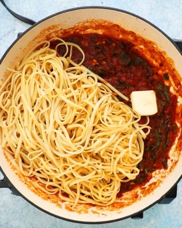 cooked fettuccini pasta along with a piece of butter and red sauce in a large white skillet.