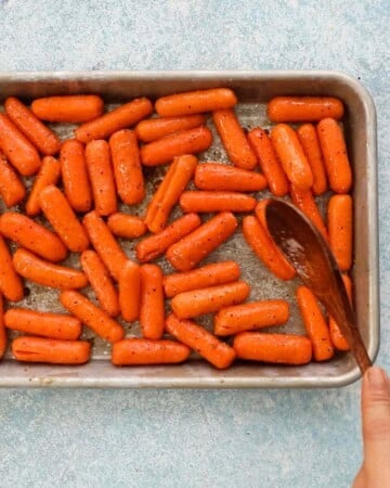 a hand mixing baby carrots in a metal sheet pan using a wooden spoon.