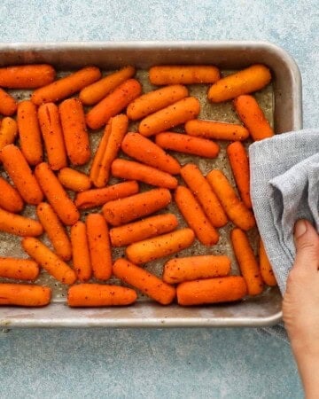 a hand holding a hot metal sheet pan with baby carrots using a grey kitchen towel.