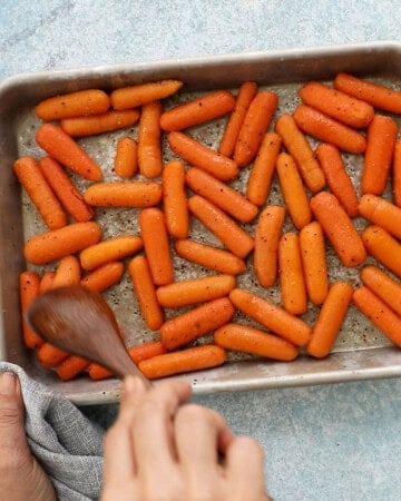 a hand mixing baby carrots in a metal sheet pan using a wooden spoon.