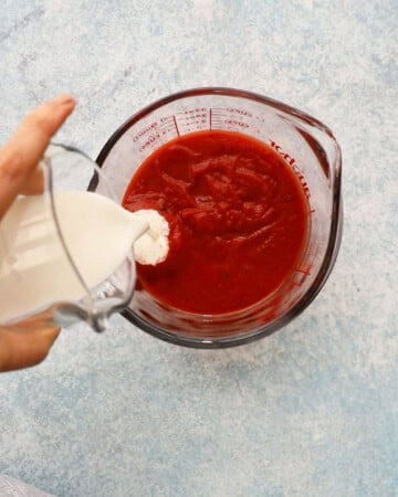 a hand pouring white cream into a bowl with red sauce.