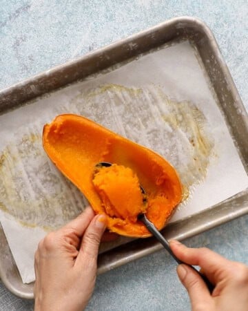 two hand scooping out the flesh from a cooked butternut squash half.