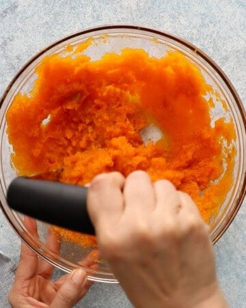 two hands mashing orange butternut squash puree in a glass bowl.