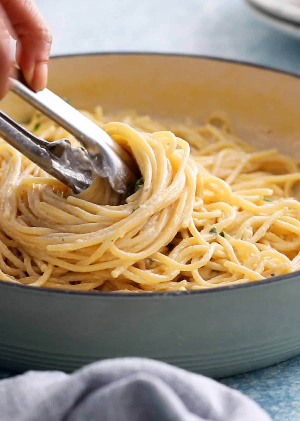 a hand lifting cooked white noodles using metal tongs from a white pan.