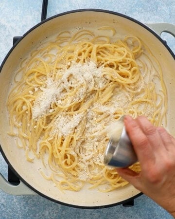 a hand adding white cheese over cooked spaghetti in white sauce in a large white pan.