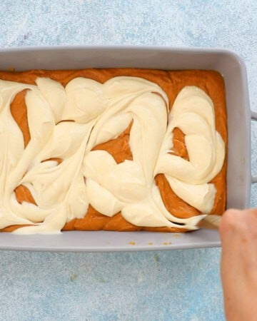 a hand swirling white cream cheese mixture with orange pumpkin cake batter in a grey pan.