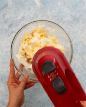 two hands beating cream cheese mixture in a glass bowl using a red electric beater.